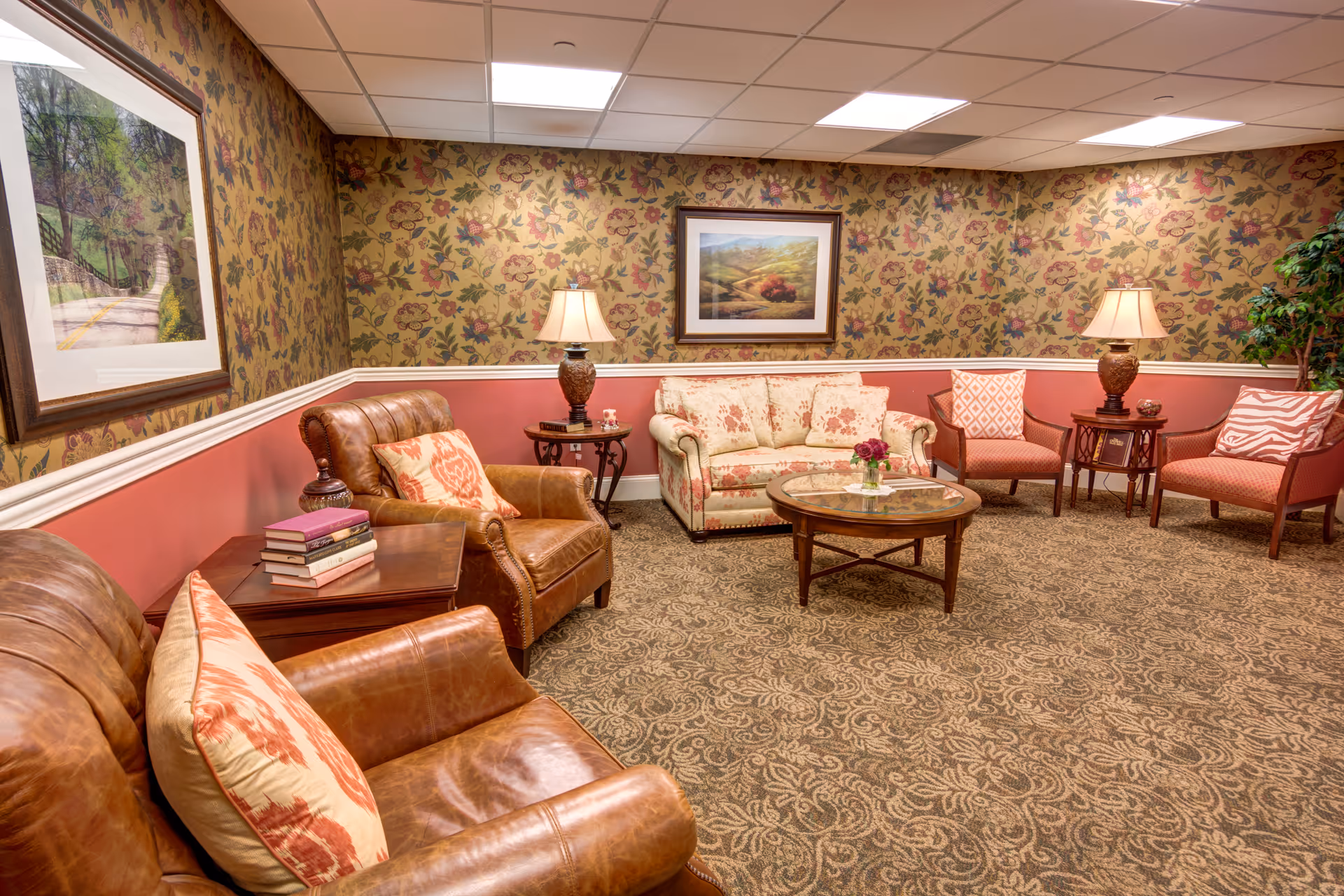 A cozy living room with floral wallpaper and a patterned carpet. The room features a floral-patterned sofa, two brown leather armchairs with decorative pillows, two coral-colored chairs with patterned cushions, two side tables with lamps, a round glass coffee table with a small flower arrangement, and framed artwork on the walls.