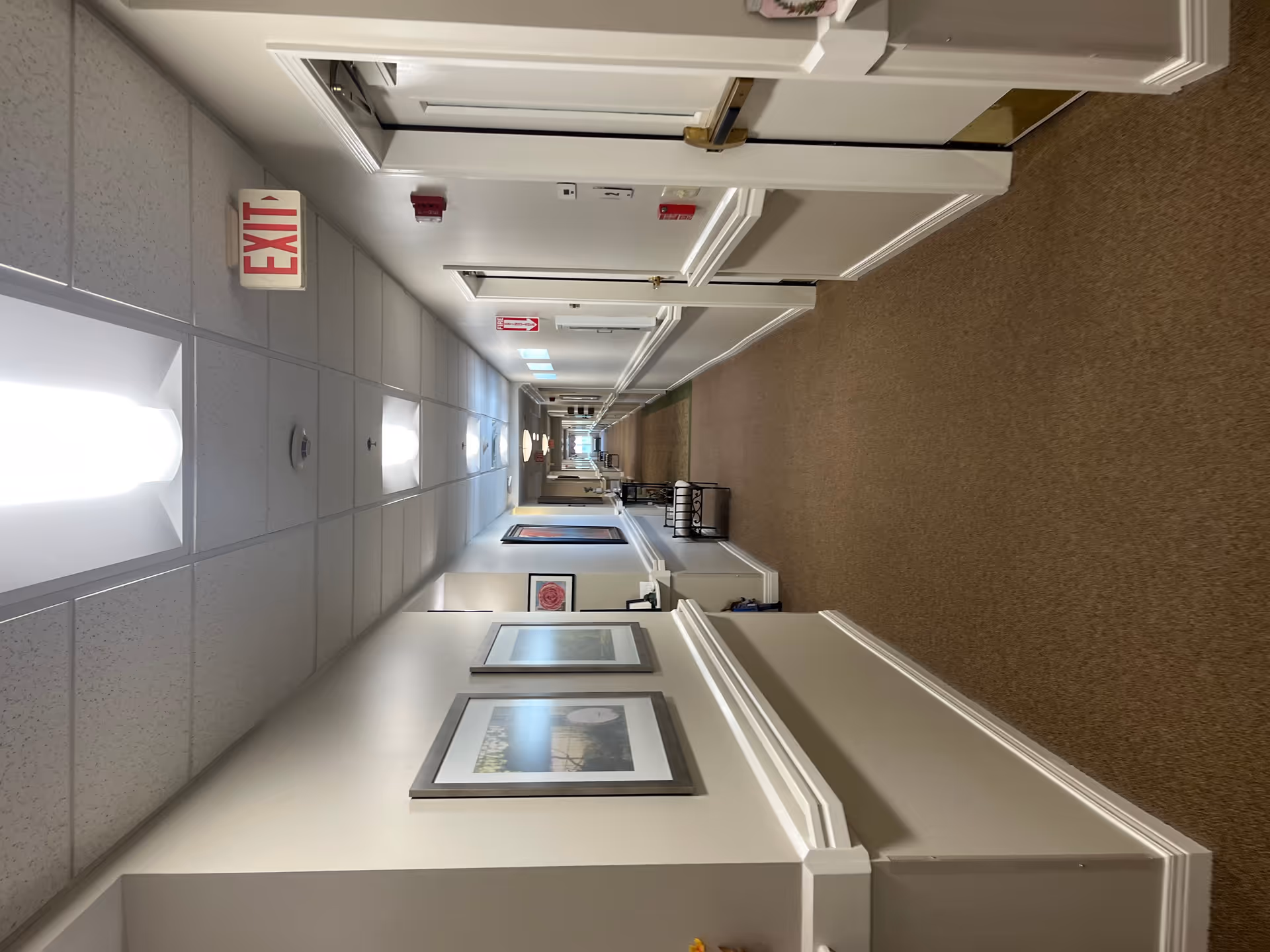 Long carpeted interior hallway in a senior living facility lined with doors, framed artwork, benches, and an EXIT sign.