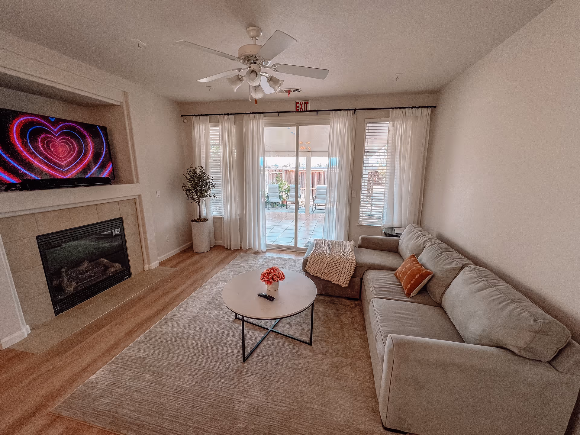 A cozy living room with a beige sectional sofa adorned with a brown pillow and a knitted throw. A round coffee table with a small flower arrangement and a remote control sits on a large beige rug. A fireplace with a mounted TV above it displaying a heart pattern is on the left wall. Sheer white curtains cover sliding glass doors that lead to an outdoor patio with chairs. A ceiling fan with lights is mounted on the ceiling.