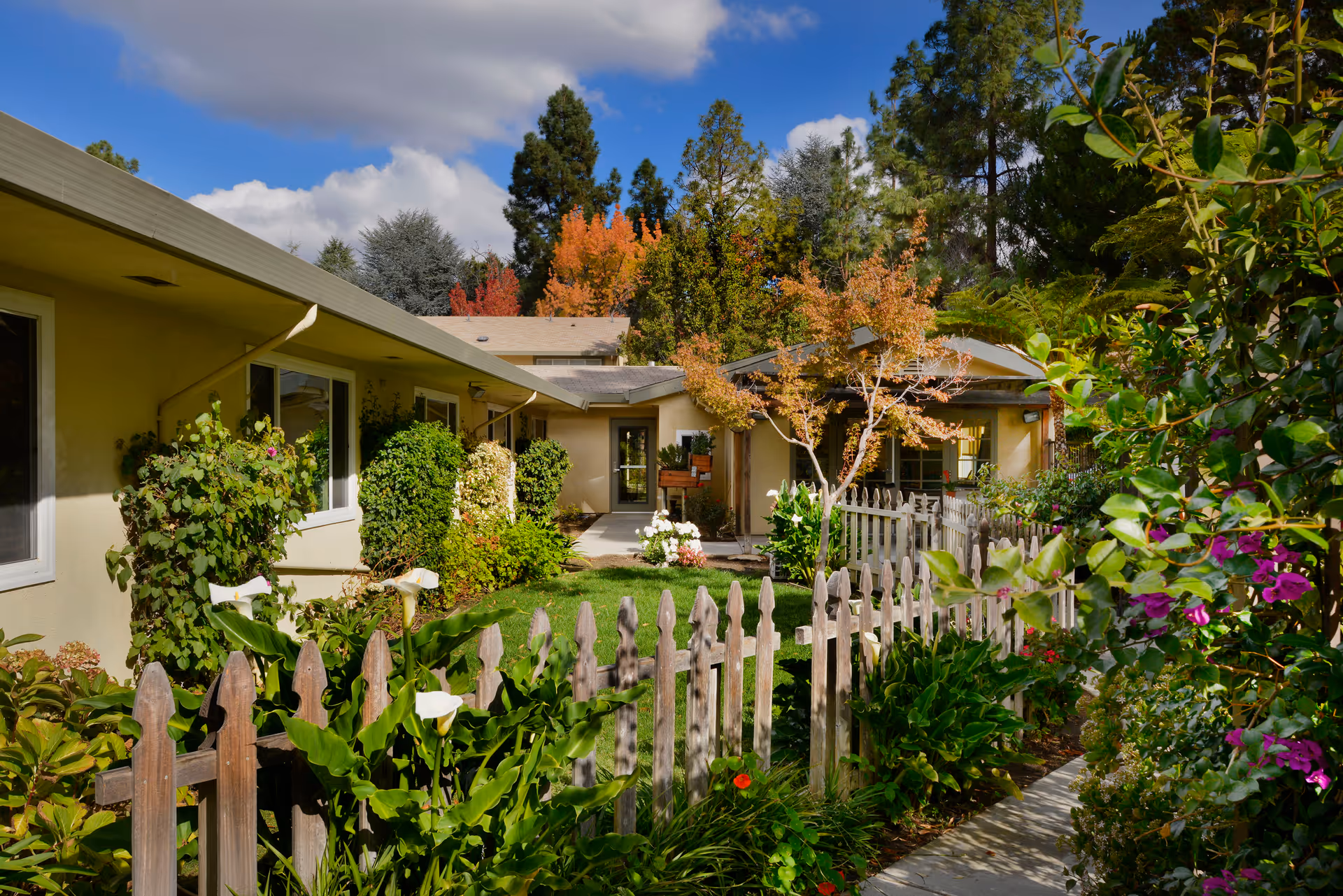 A sunny outdoor garden area at a senior living facility with a wooden picket fence, lush green plants, colorful flowers, and trees with autumn foliage. The building exterior is light-colored with windows and a covered walkway leading to an entrance.