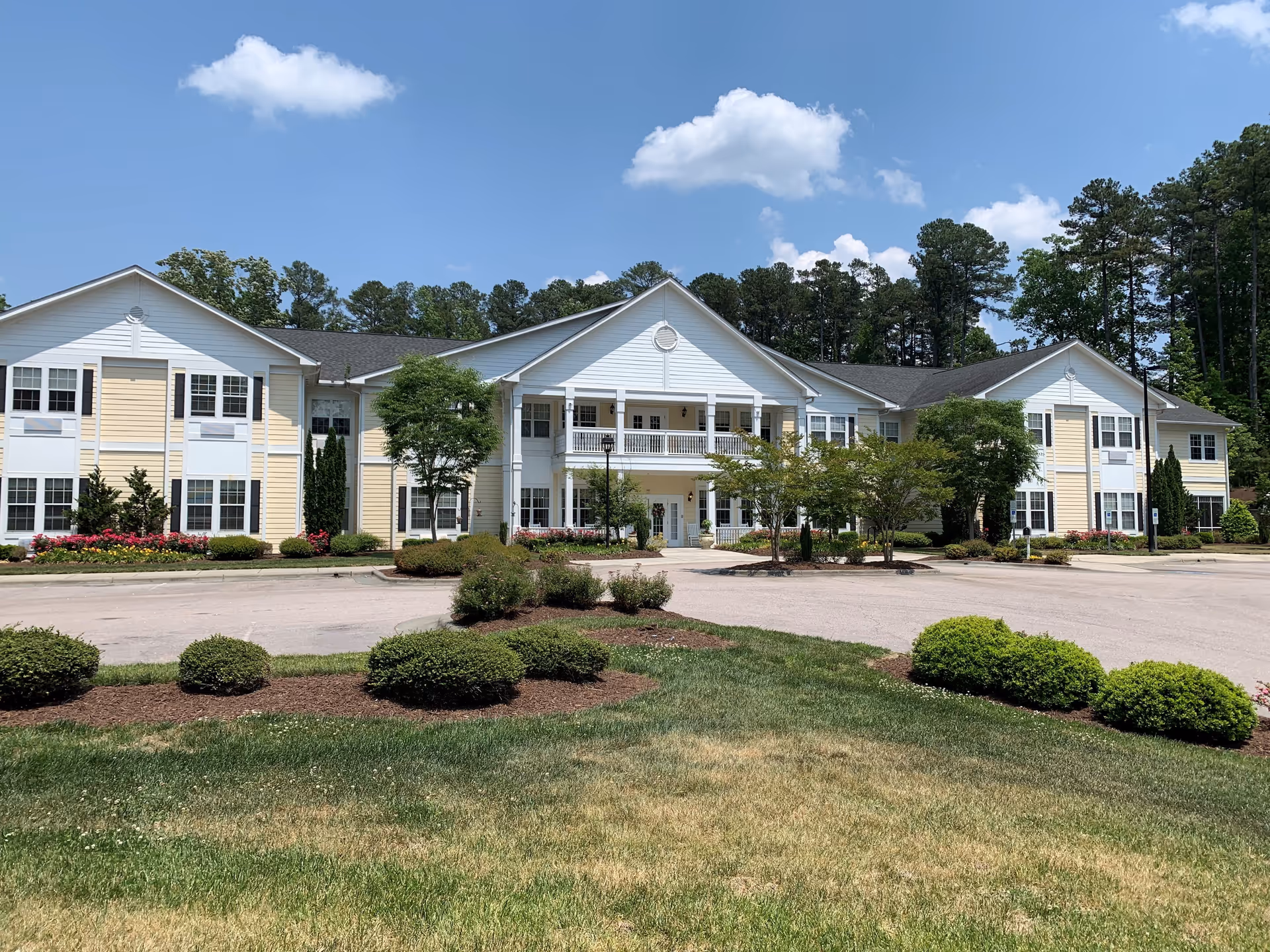 Exterior view of TerraBella Durham, a two-story senior living facility with white and light yellow siding, surrounded by manicured bushes, trees, and a green lawn under a blue sky with some clouds.