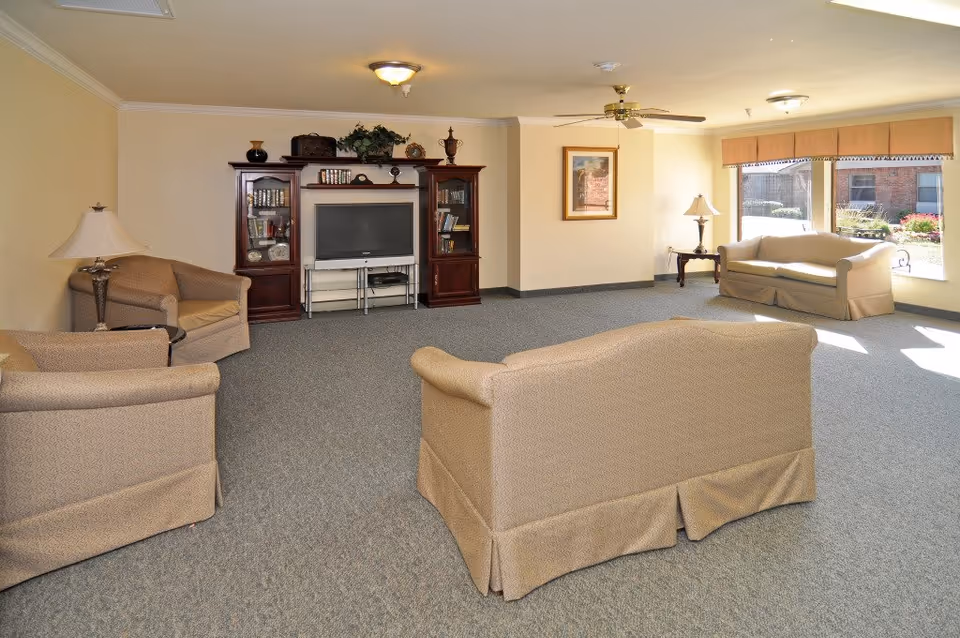 A spacious living room with beige upholstered sofas and armchairs arranged around a wooden entertainment center with a flat-screen TV. The room has carpeted flooring, cream-colored walls, ceiling lights, a ceiling fan, and large windows letting in natural light. There are decorative items on the entertainment center and a framed picture on the wall.