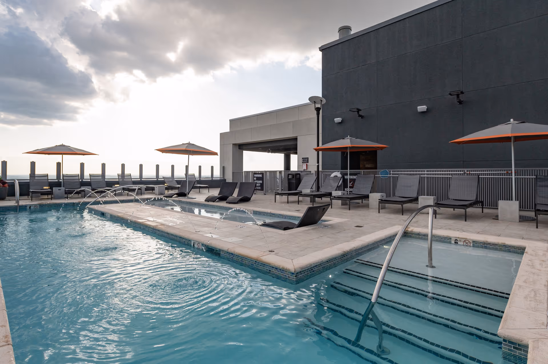 Outdoor swimming pool area with lounge chairs and umbrellas on a rooftop, with water fountains arching into the pool and a cloudy sky in the background.