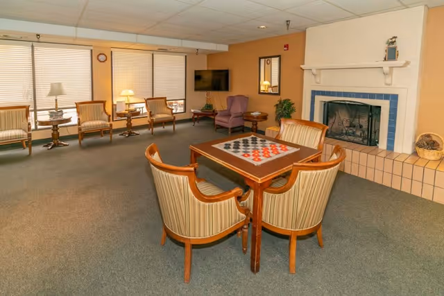 Communal lounge with a game table and four chairs in the foreground, additional seating, TV, lamps, and a fireplace along the walls.