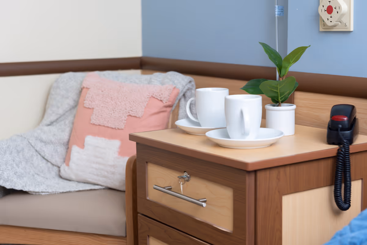 A cozy corner with a cushioned bench covered by a gray blanket and a pink and white decorative pillow. Next to the bench is a wooden nightstand with two white cups on saucers, a small potted plant, and a black telephone with a coiled cord. The wall behind is painted blue and beige with a brown trim.
