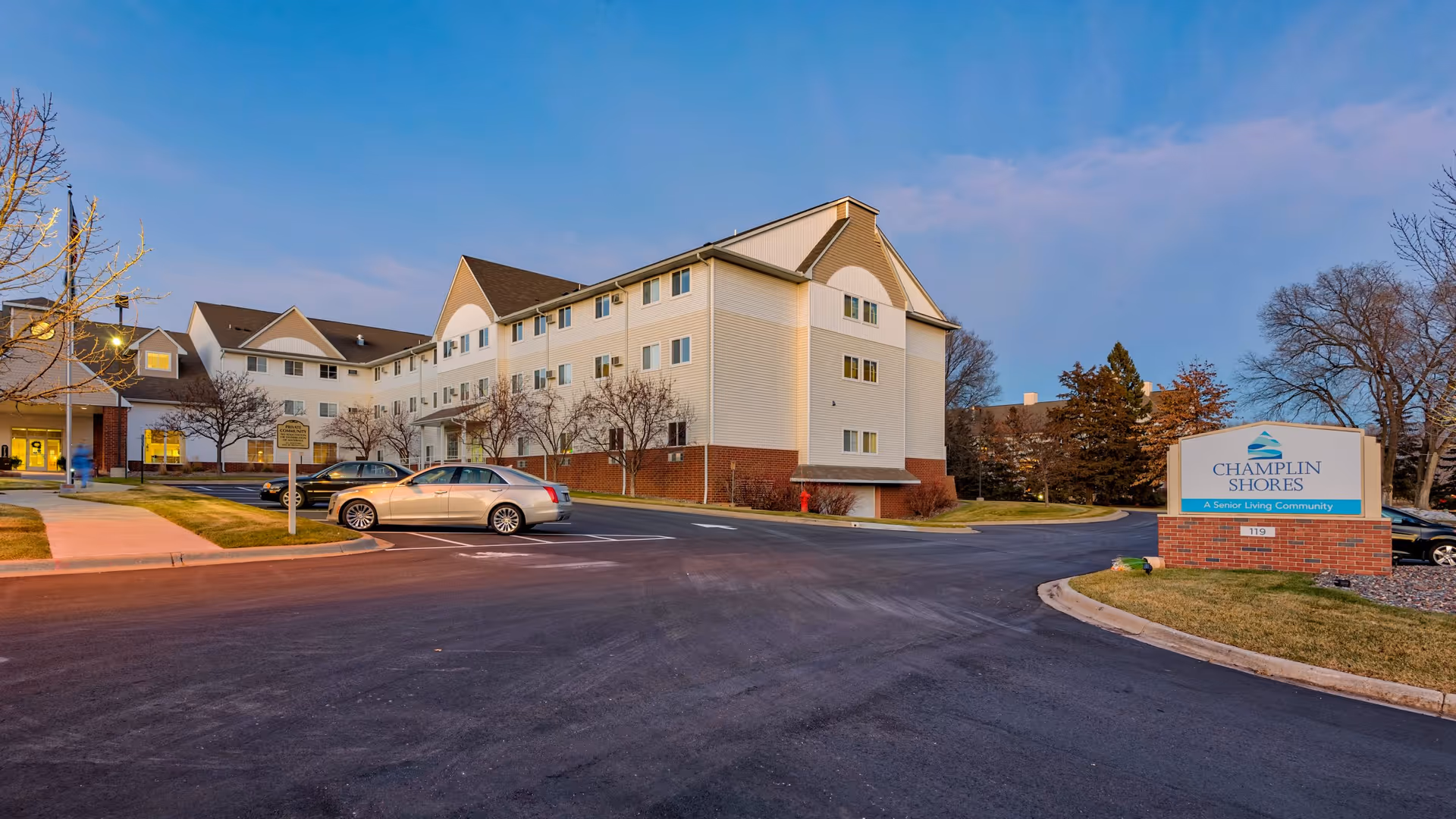 Exterior view of a multi-story senior living building and parking lot at dusk with a Champlin Shores sign in the foreground.