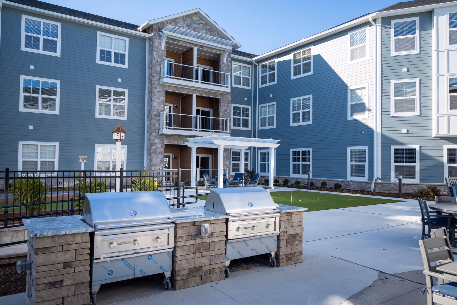 Outdoor courtyard area of a senior living facility with two stainless steel built-in grills, patio seating, a small green lawn, and a three-story building with balconies and multiple windows in the background.