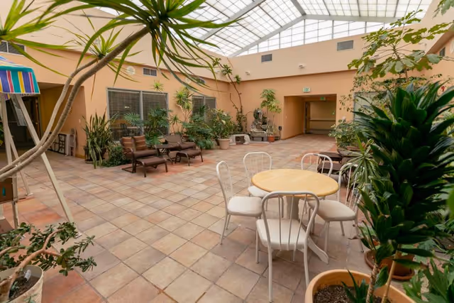 Indoor courtyard area with a tiled floor and a glass ceiling allowing natural light. There are several potted plants and small trees around the space. In the foreground, there is a round table with four white chairs. In the background, there are two brown lounge chairs and more plants near the walls. The walls are painted a light peach color.