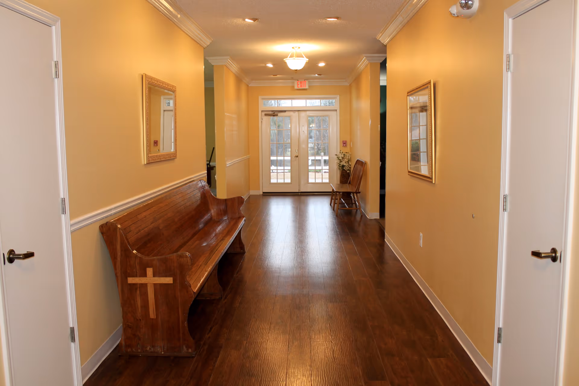A well-lit hallway with wooden flooring and yellow walls. There are two white doors on each side of the hallway, a wooden bench with a cross carved on the side on the left, and two wooden chairs with a vase of flowers on the right near the glass double doors at the end. Two framed mirrors hang on the walls, and ceiling lights illuminate the space.