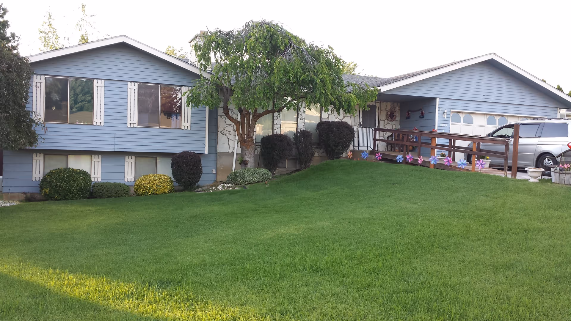 Exterior view of a single-story blue house with white trim, a green lawn, various bushes, a tree in front, a wooden ramp decorated with colorful pinwheels, and a silver minivan parked in the driveway.