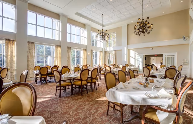 Bright dining room with round tables set with white tablecloths and upholstered chairs beneath chandeliers and large windows.