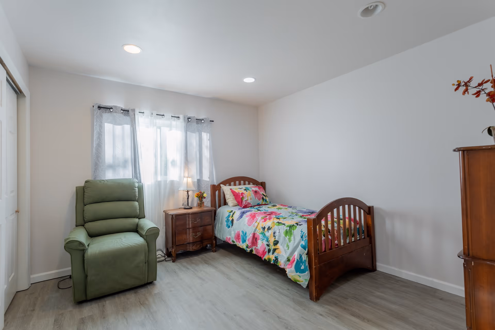 A bright bedroom with a single wooden bed covered in a colorful floral bedspread, a green recliner chair, a wooden nightstand with a lamp and flowers, and a wooden dresser. The room has light gray walls, wood flooring, and a window with sheer and gray curtains.