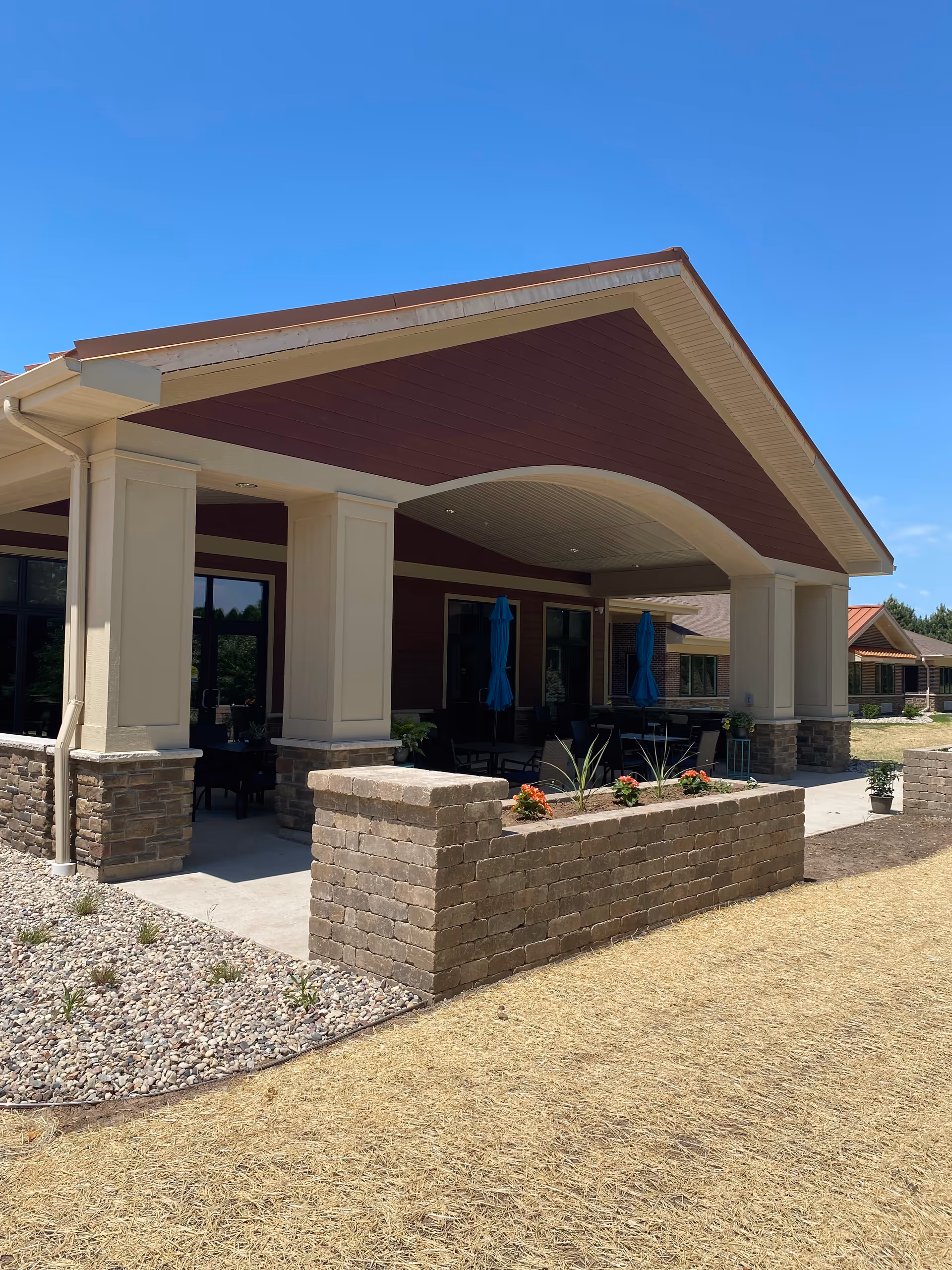 Covered outdoor patio area with stone pillars and brick planter boxes containing flowers, attached to a building with red siding and beige trim under a clear blue sky.