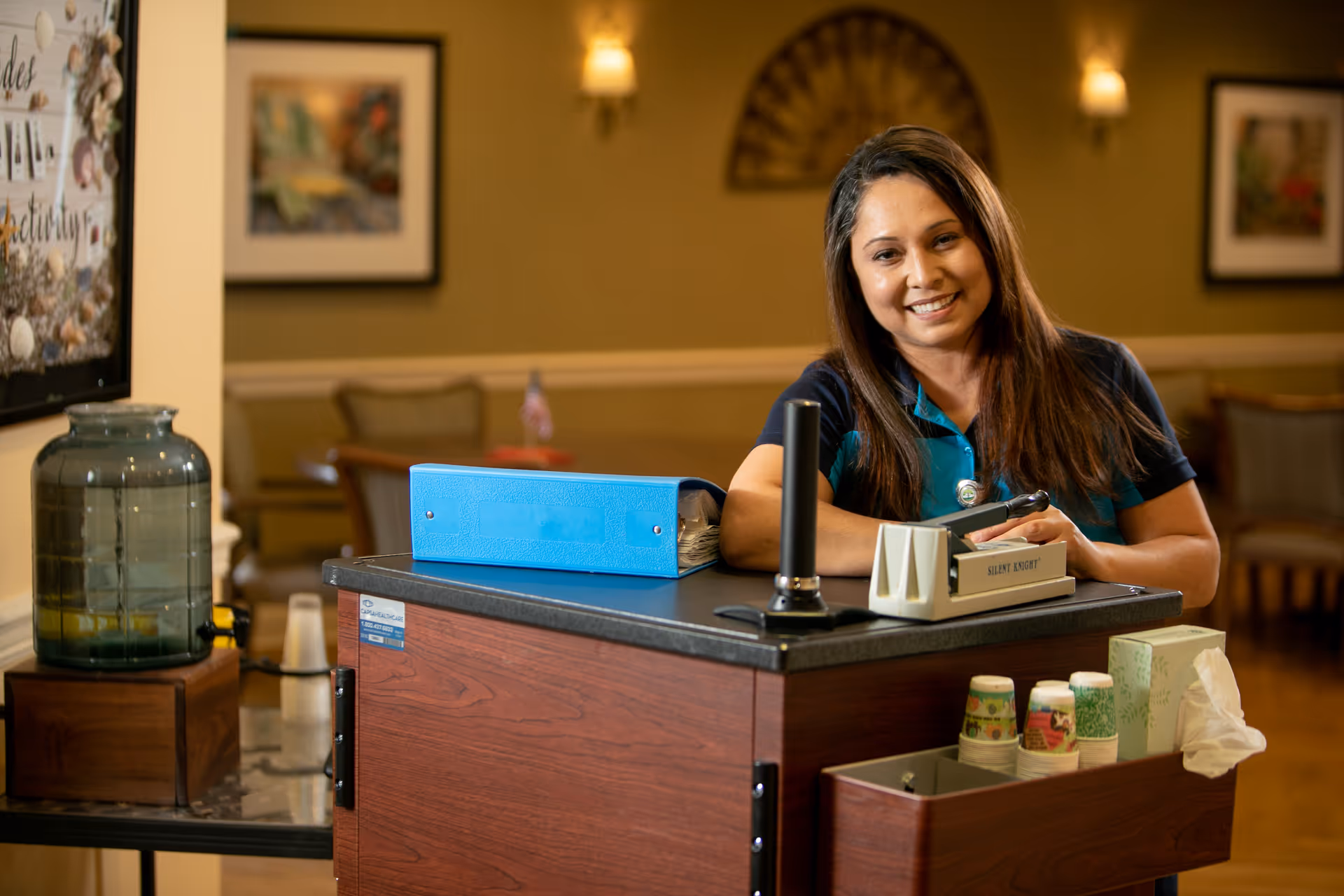 A smiling woman with long dark hair wearing a blue and teal uniform leans on a wooden cart with office supplies including a blue binder and a stapler. The background shows a warmly lit room with chairs, framed pictures on the wall, and a water dispenser on a small table.