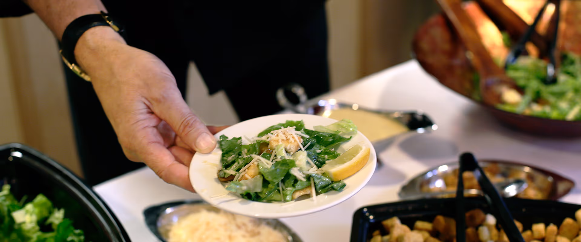 A hand holding a small plate with a serving of Caesar salad, including romaine lettuce, grated cheese, croutons, and a lemon wedge, with salad serving bowls and utensils visible on a table in the background.