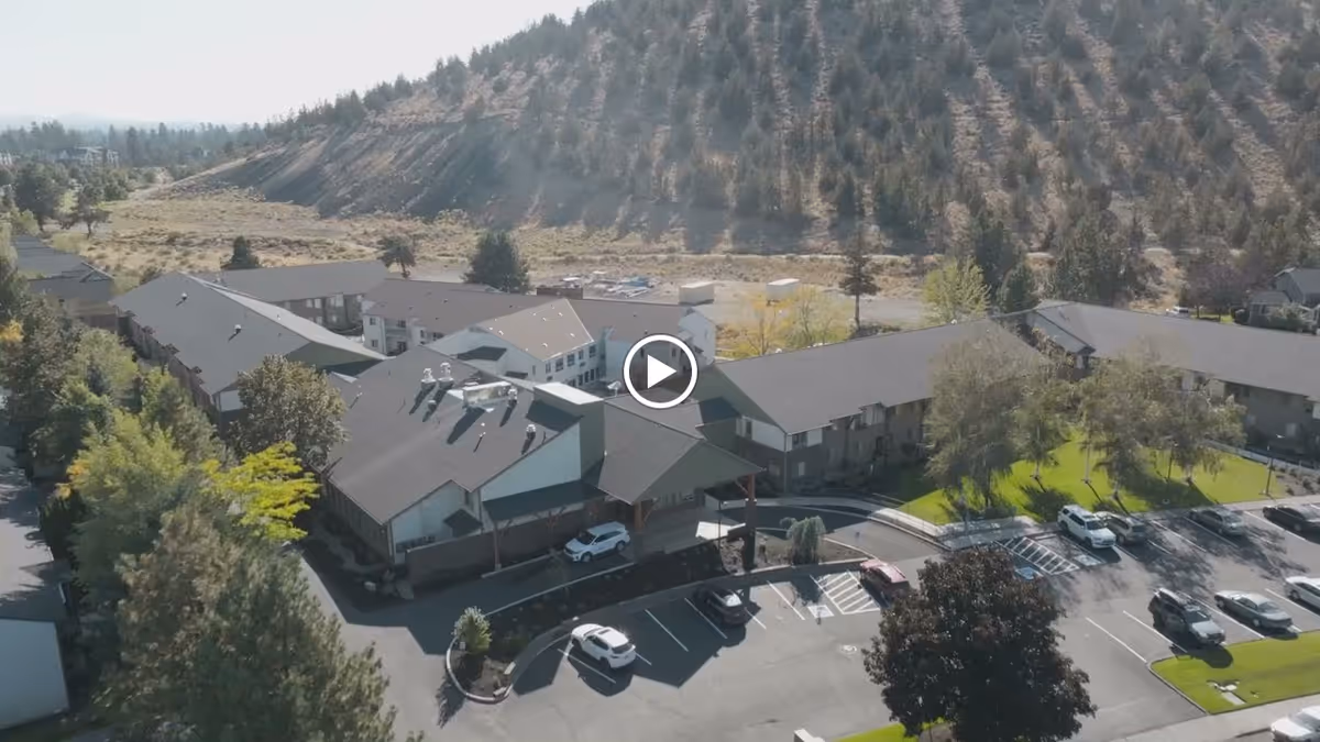 Aerial view of Cascades of Bend Retirement Community showing multiple connected buildings with dark roofs, surrounded by trees and parking lots. The facility is located near a hillside with sparse vegetation.