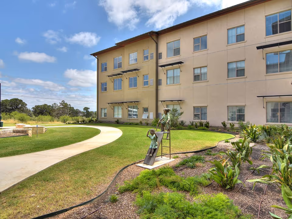 Outdoor garden area at Élan Westpointe at New Braunfels featuring a curved concrete pathway, green grass, landscaped plants, and a bronze sculpture of two children playing on a slide in front of a three-story beige building under a partly cloudy sky.