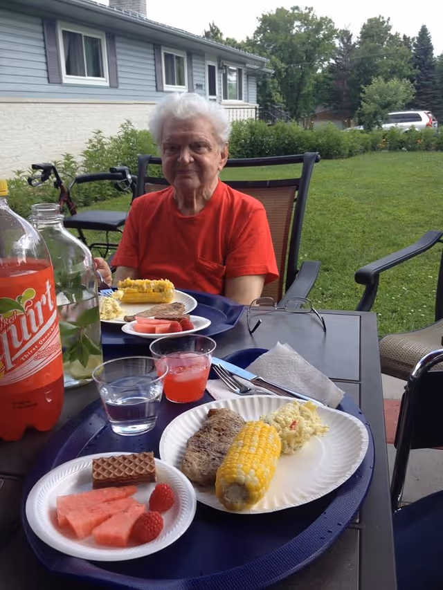 An elderly person wearing a red shirt sitting at an outdoor table with plates of food including corn on the cob, watermelon, raspberries, a grilled patty, and a side salad. There are drinks on the table and a house with greenery in the background.