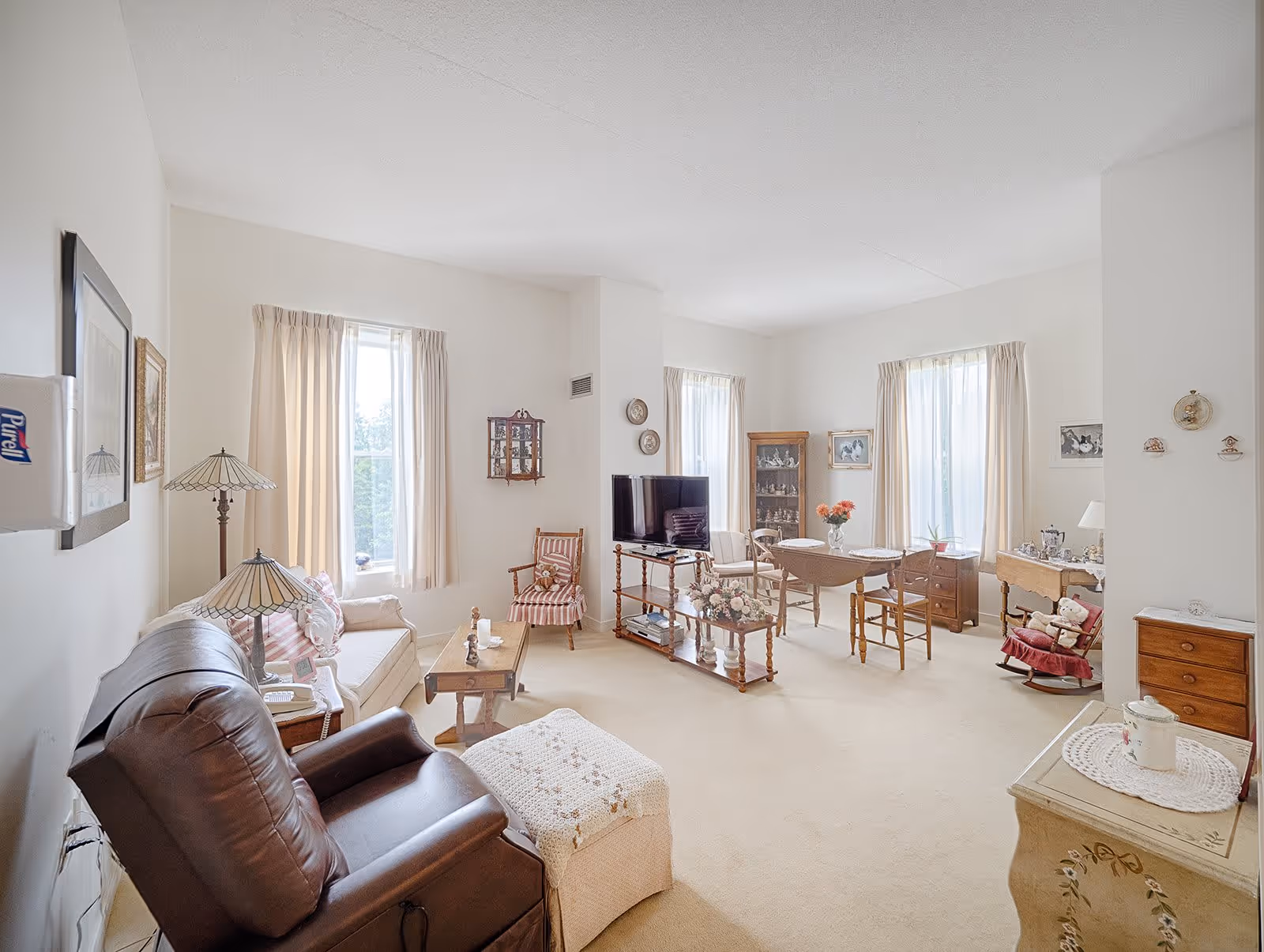 A bright and cozy living room with beige walls and carpet. The room features a brown leather recliner, a beige sofa with striped cushions, a wooden coffee table, and a TV on a wooden stand. There are three windows with light curtains allowing natural light to fill the space. Additional furniture includes a wooden dining table with chairs, a display cabinet, and various decorative items such as lamps, framed pictures, and flowers.