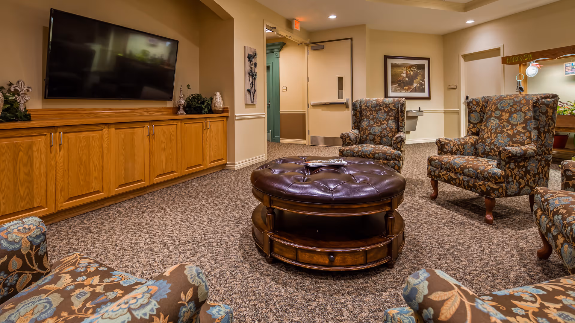 A cozy living room area with patterned armchairs arranged around a round, tufted leather ottoman. There is a large flat-screen TV mounted on a wooden cabinet along one wall, and a framed painting hangs on another wall. The room has carpeted flooring and beige walls with white trim.