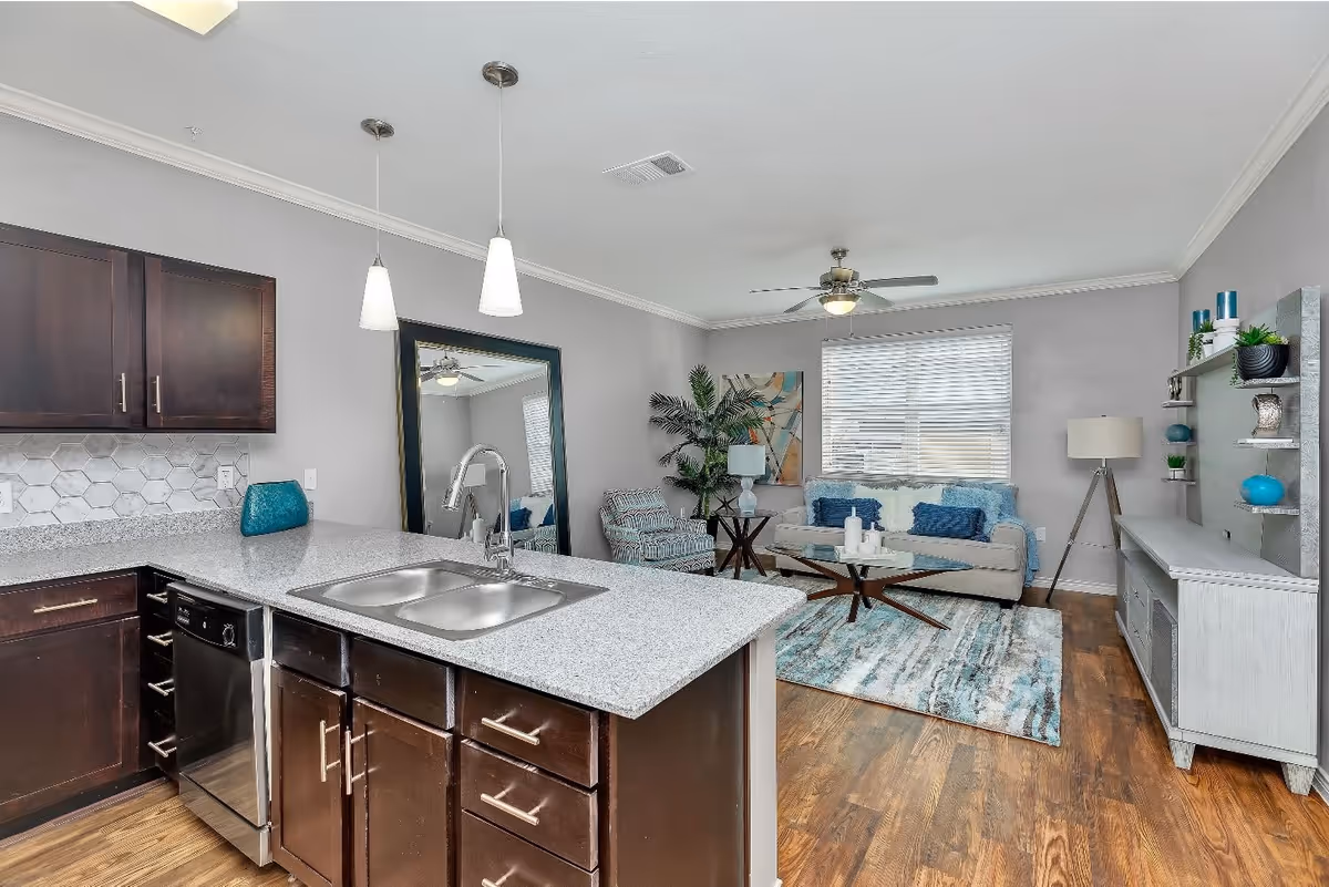 Open-plan interior showing a kitchen island with a sink in the foreground and a living area with a sofa, coffee table, rug, and TV console in the background.