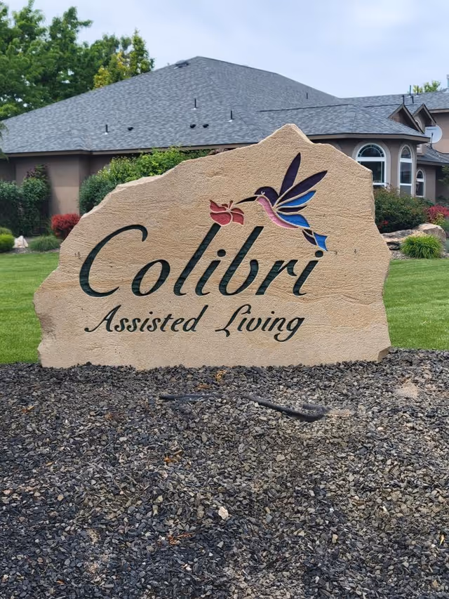 A large stone sign with the text 'Colibri Assisted Living' and an image of a colorful hummingbird holding a flower, set in front of a residential building with a gray roof and green landscaping.