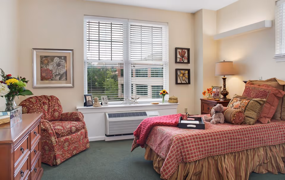 Cozy bedroom with a patterned bed and pillows (plus a teddy bear), an upholstered armchair, dresser, bedside table with lamp, and a large window letting in daylight.