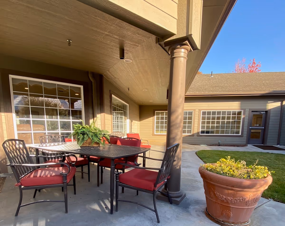 Outdoor patio area at Summer Wood Alzheimer's Special Care with a black metal table and six chairs with red cushions. There is a large potted plant on the table and another large terracotta planter with greenery on the concrete floor. The patio is covered with a ceiling supported by a column, and the building has large windows and a door leading inside. The sky is clear and blue.