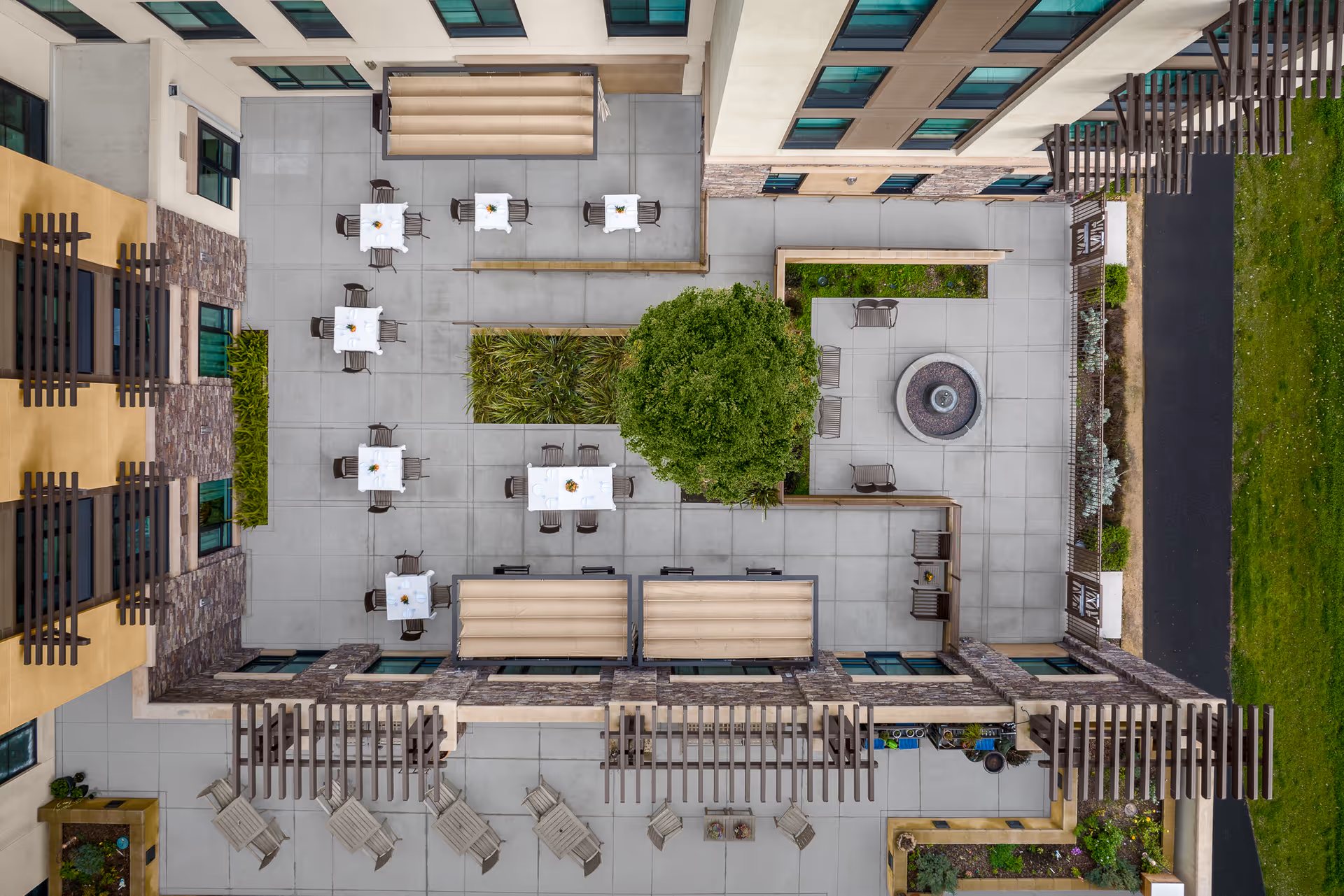 Aerial view of an outdoor courtyard at Belmont Village Senior Living Albany featuring multiple tables with chairs, a central tree surrounded by greenery, a circular fire pit with seating, and pergolas providing partial shade along the building's exterior.