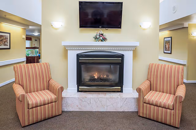 A cozy interior space featuring a lit fireplace with a white mantel, a floral arrangement on top, and a flat-screen TV mounted above. Two striped armchairs are positioned on either side of the fireplace. The walls are painted a soft yellow, and the carpet is brown. There are doorways visible on both sides leading to other rooms.