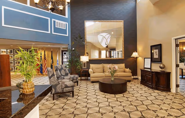 A spacious and well-lit living room area with a beige sofa, two patterned armchairs, a round dark wooden coffee table with a vase of flowers, a large decorative mirror on a dark accent wall, and a wooden sideboard with a lamp and framed picture. The floor is covered with a patterned carpet, and there are plants and flags visible in the background.