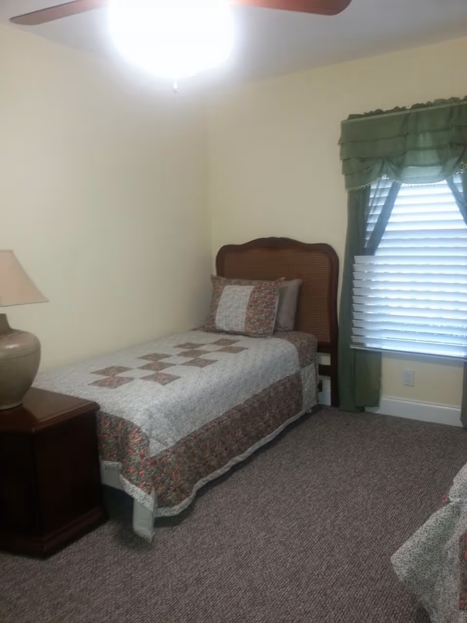 A small bedroom with a single bed covered with a patterned quilt and matching pillows. There is a wooden nightstand with a lamp on it next to the bed. A window with green curtains and white blinds is on the right side of the room. The walls are light-colored and the floor is carpeted.