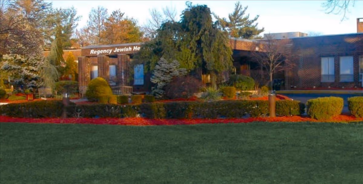 Exterior view of Regency Jewish Heritage Nursing and Post-Acute Rehabilitation Center showing a brick building surrounded by landscaped bushes, trees, and a well-maintained lawn under a clear sky.