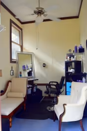 A small room with cream-colored walls and a ceiling fan. The room contains two upholstered chairs, a black salon chair in front of a mirror, and a shelving unit with various hair and beauty products. There is a window on the left wall letting in natural light.