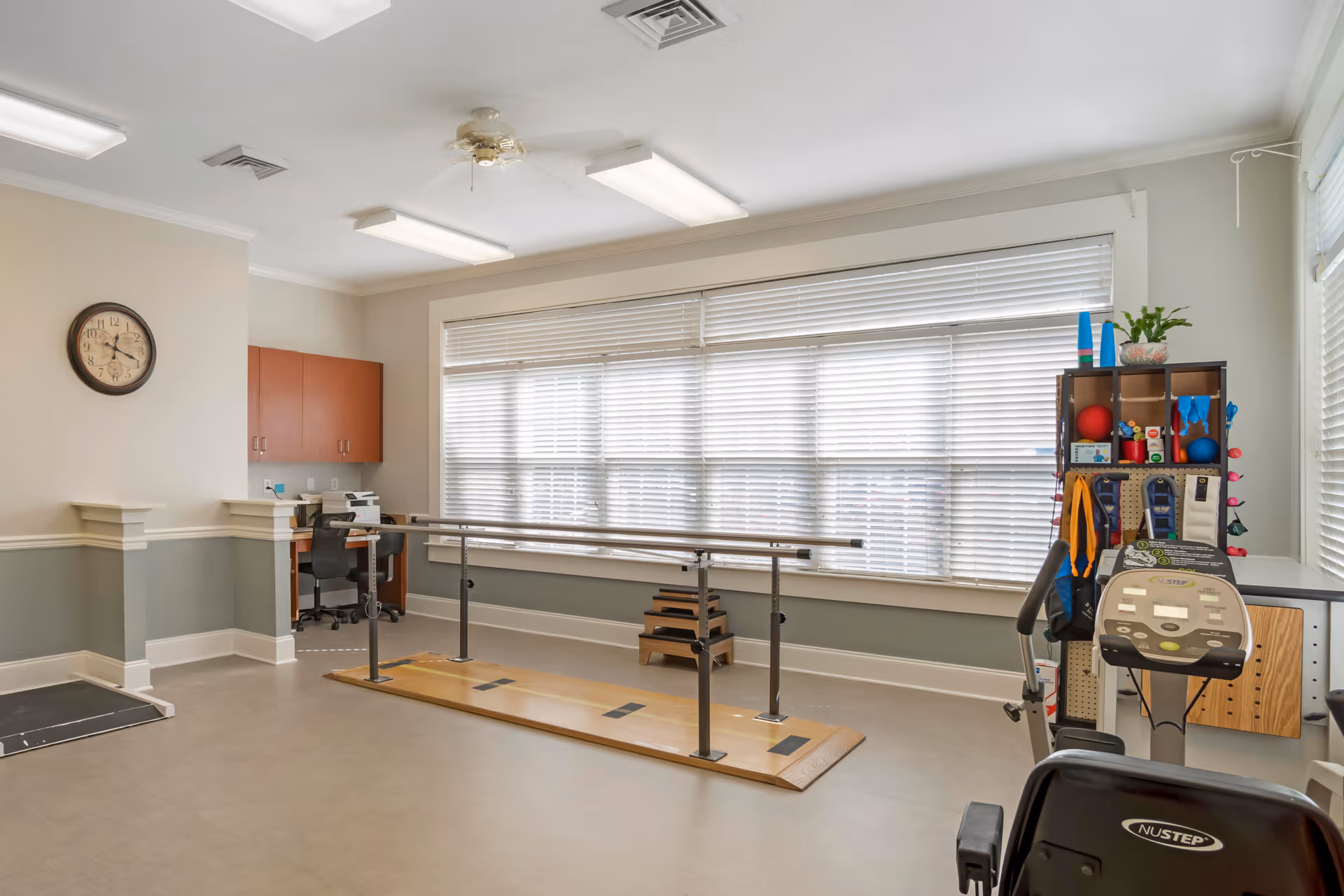 A rehabilitation or physical therapy room with parallel bars for walking exercises, a stationary exercise bike, a small desk with a chair and printer, and shelves holding various therapy equipment and supplies. The room has large windows with blinds, a wall clock, and overhead fluorescent lighting.