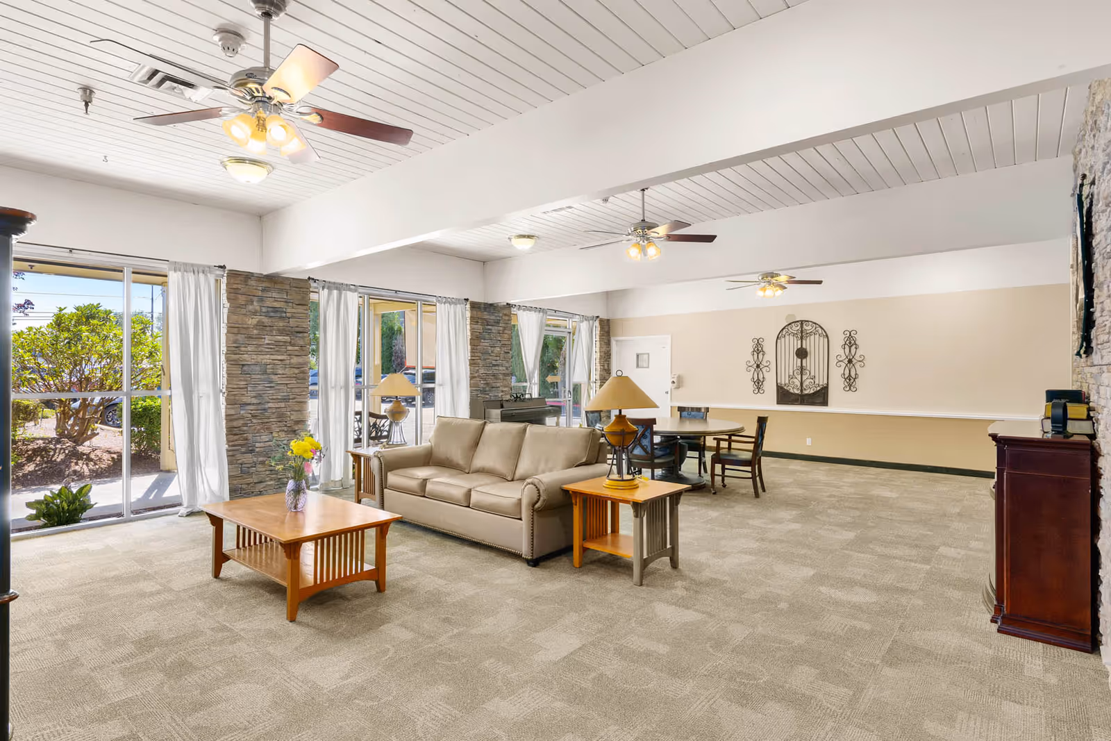 Spacious bright common lounge with a sofa, coffee and side tables, lamps, dining area, ceiling fans, large windows and stone-accent columns.