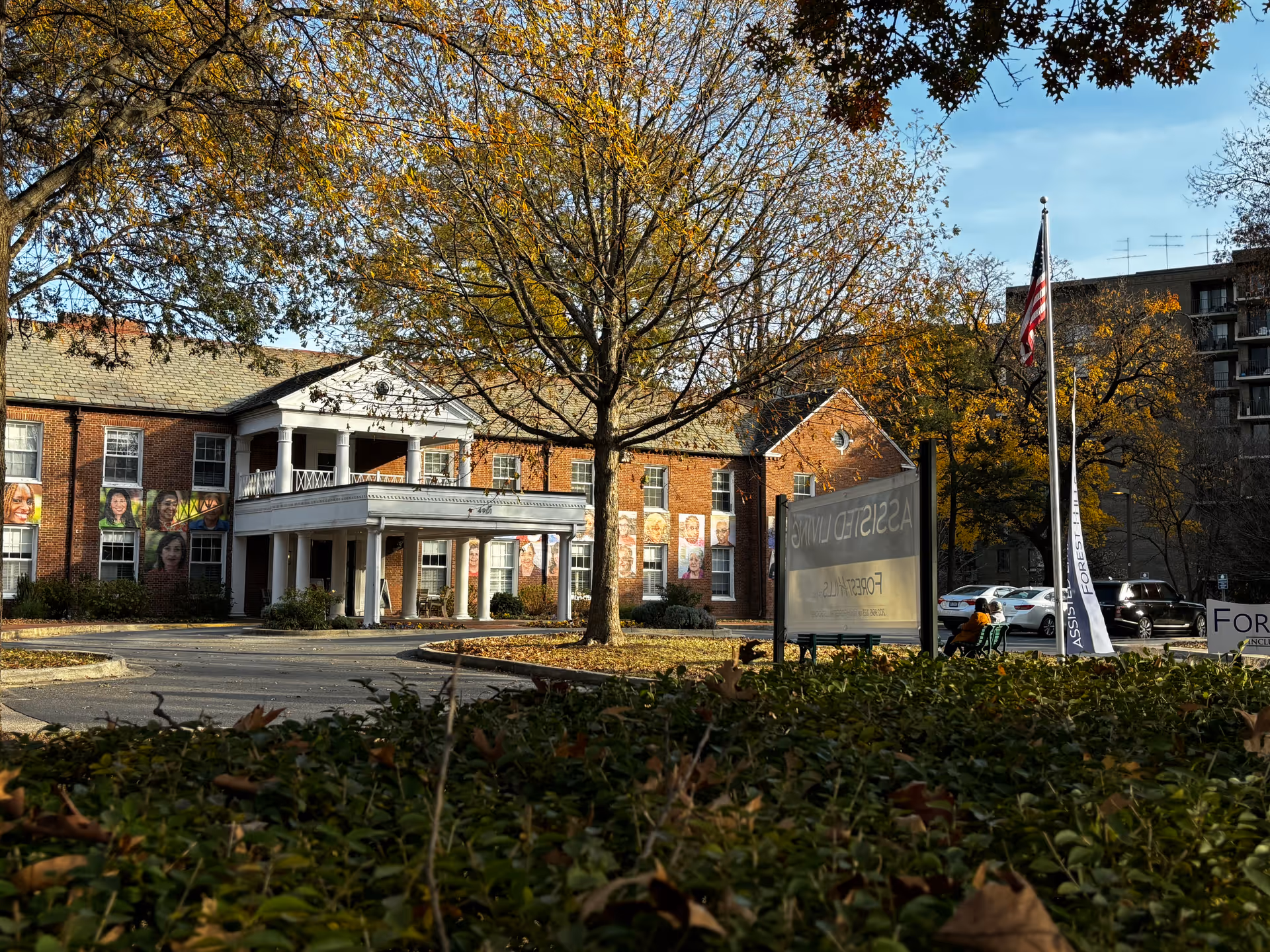 Exterior view of a brick building with white columns and a covered entrance, surrounded by trees with autumn leaves. There is a flagpole with an American flag and a sign partially visible that reads 'ASSISTED LIVING' and 'FOREST SIDE'. Cars are parked nearby and a person is sitting on a bench near the sign.