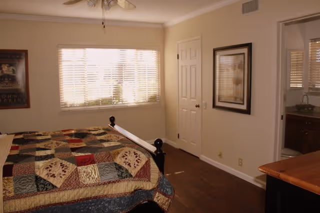 A bedroom with a bed covered in a colorful patchwork quilt, a window with blinds letting in natural light, a closed white door, a framed picture on the wall, and a view into an adjacent bathroom with a sink and mirror.