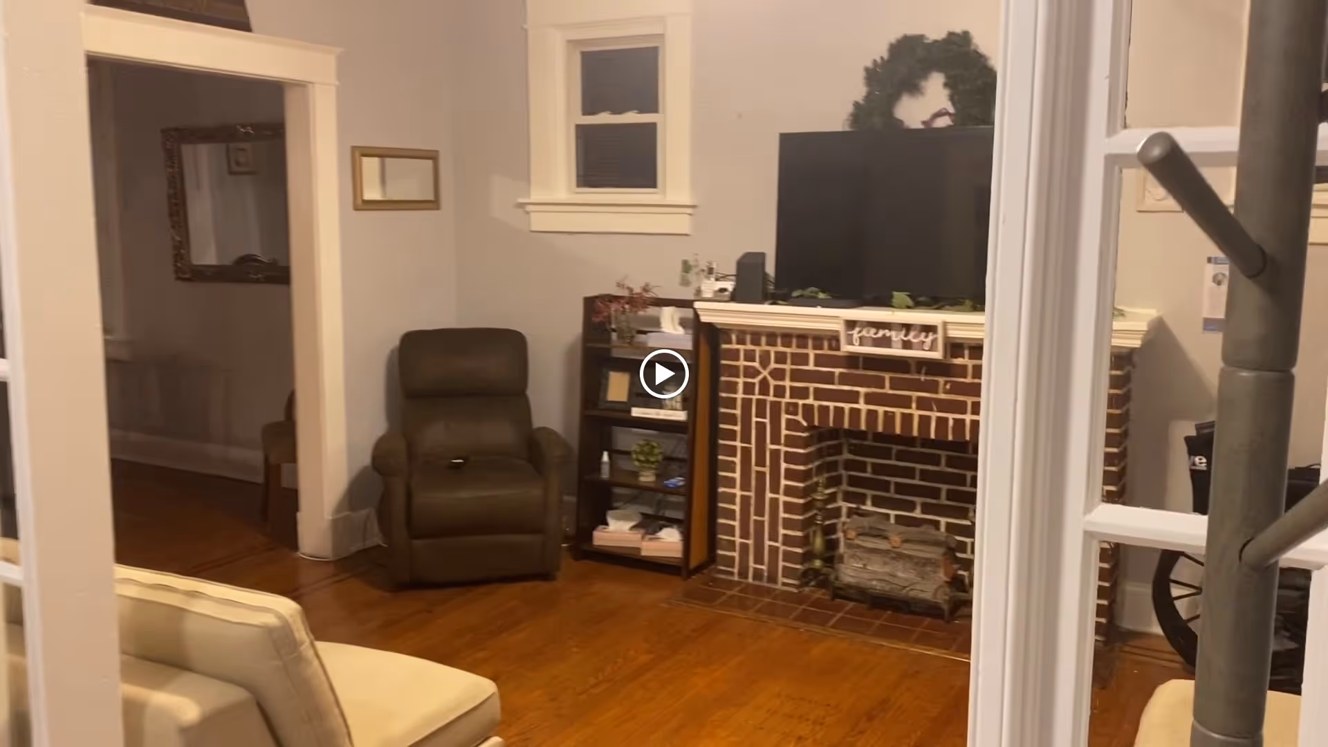 A cozy living room with wooden floors, a brown recliner chair, a beige sofa, a brick fireplace with a TV on the mantel, and a small bookshelf filled with books and decorative items.