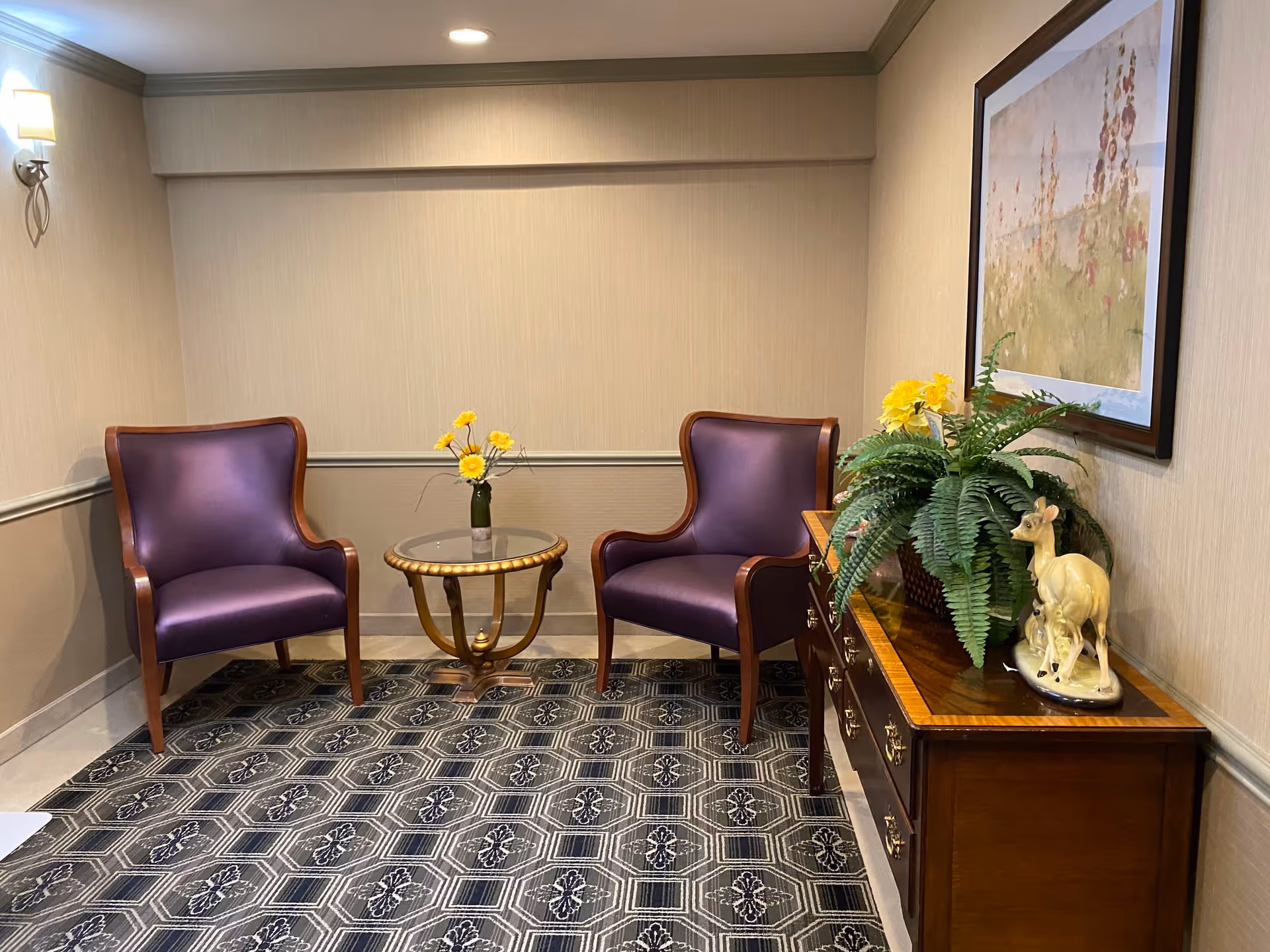 A small sitting area with two purple upholstered armchairs facing each other with a round glass-top table between them holding a vase with yellow flowers. To the right, there is a wooden dresser with a potted fern, a ceramic deer figurine, and a framed floral painting hanging above it. The room has beige walls, a patterned carpet, and soft lighting from a wall sconce and recessed ceiling light.