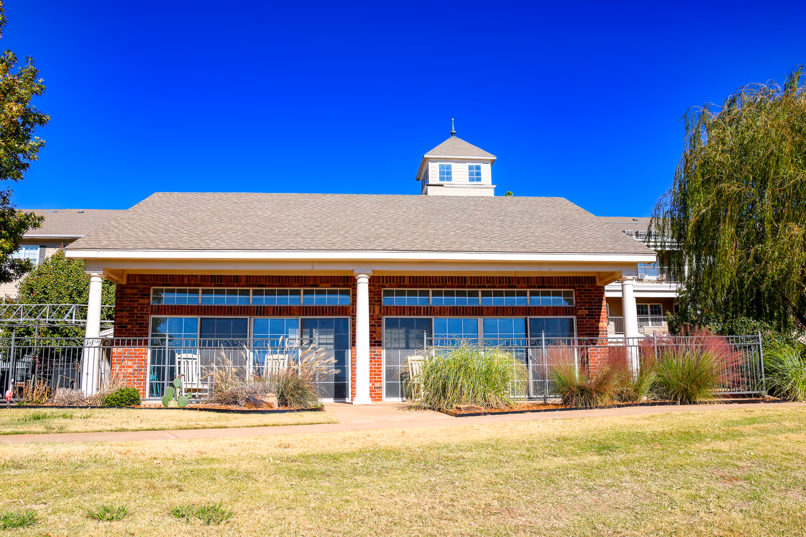 Exterior view of a single-story brick building with large windows and white columns, surrounded by ornamental grasses and a metal fence, under a clear blue sky.