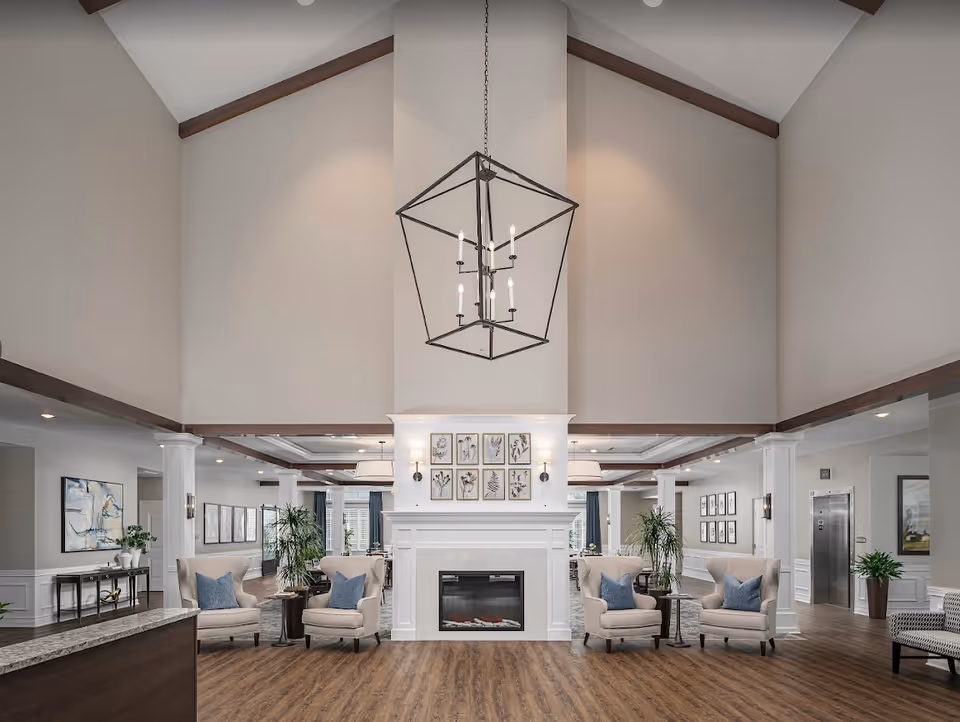 Spacious vaulted lobby with a central fireplace, chandelier, and seating areas with armchairs and plants.