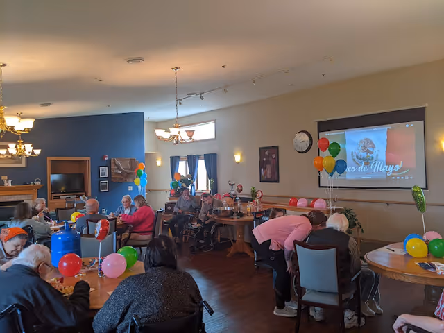 Residents seated at decorated tables in a senior living dining/activity room with balloons and a projection screen.