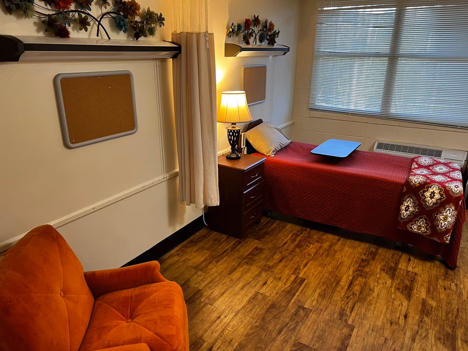 A small resident bedroom with a single bed covered in a red quilt, a bedside table and lamp, an orange armchair, and a window with blinds.