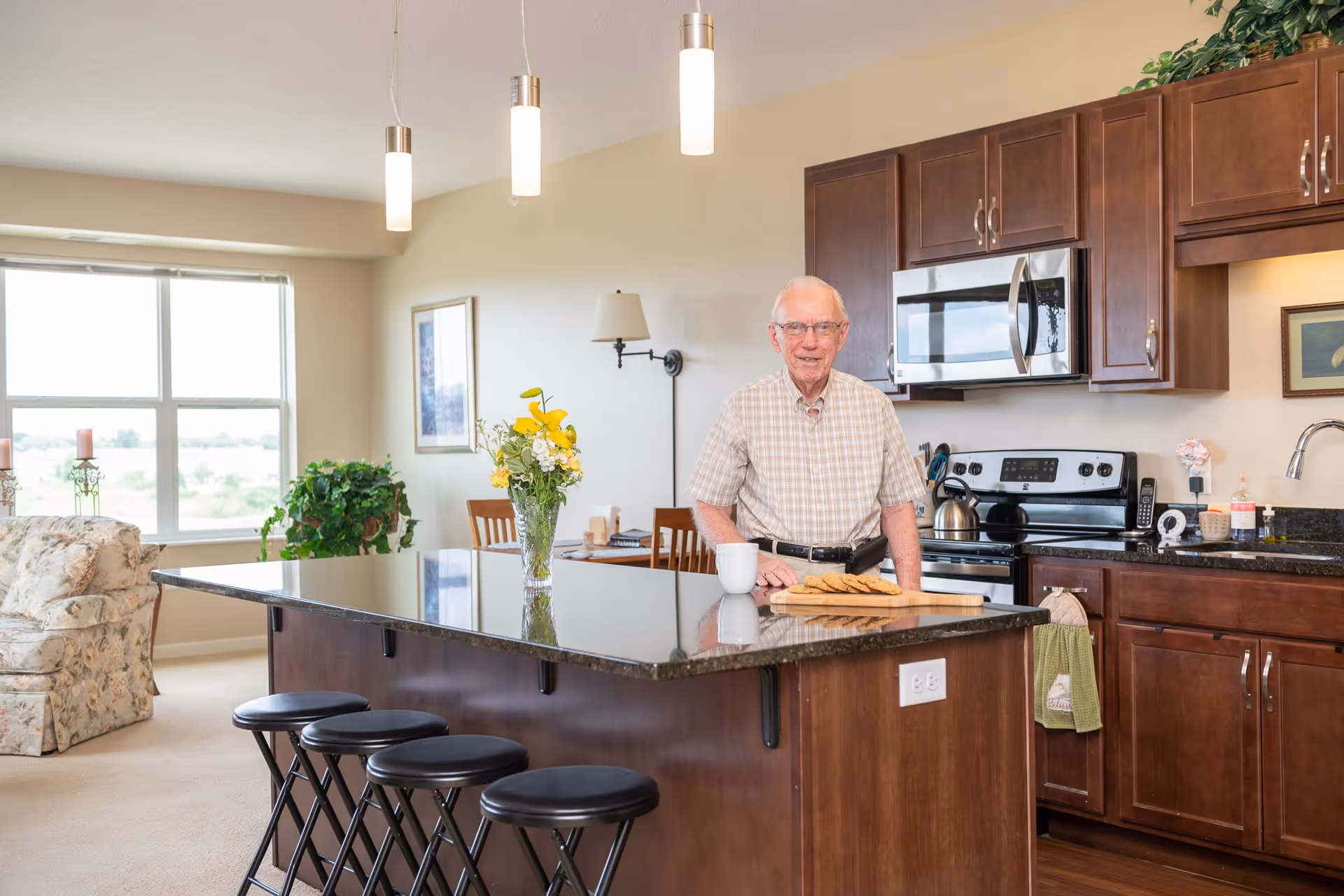 An elderly man standing behind a kitchen island in a well-lit kitchen and living area. The kitchen features dark wooden cabinets, a microwave, stove, and a granite countertop island with four black stools. A vase with yellow flowers and a plate of cookies are on the island. In the background, there is a floral armchair, a large window, and some indoor plants.