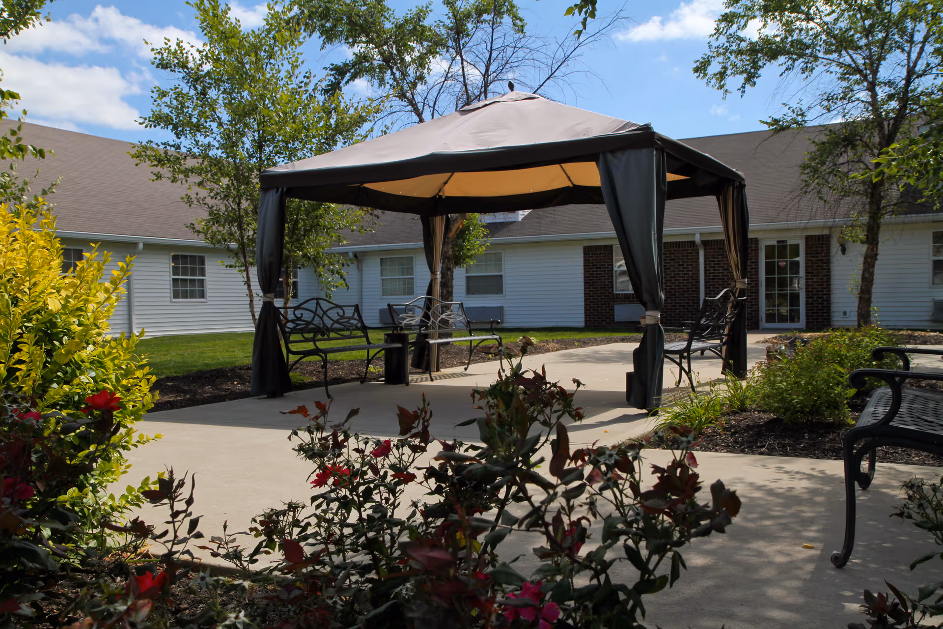 Outdoor patio area with a gazebo featuring a beige canopy and dark curtains, surrounded by benches and greenery including bushes and trees, with a white building in the background under a partly cloudy sky.