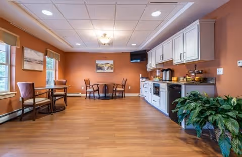 Communal dining and kitchenette area with small tables, chairs, cabinets, and a TV in a senior living facility.