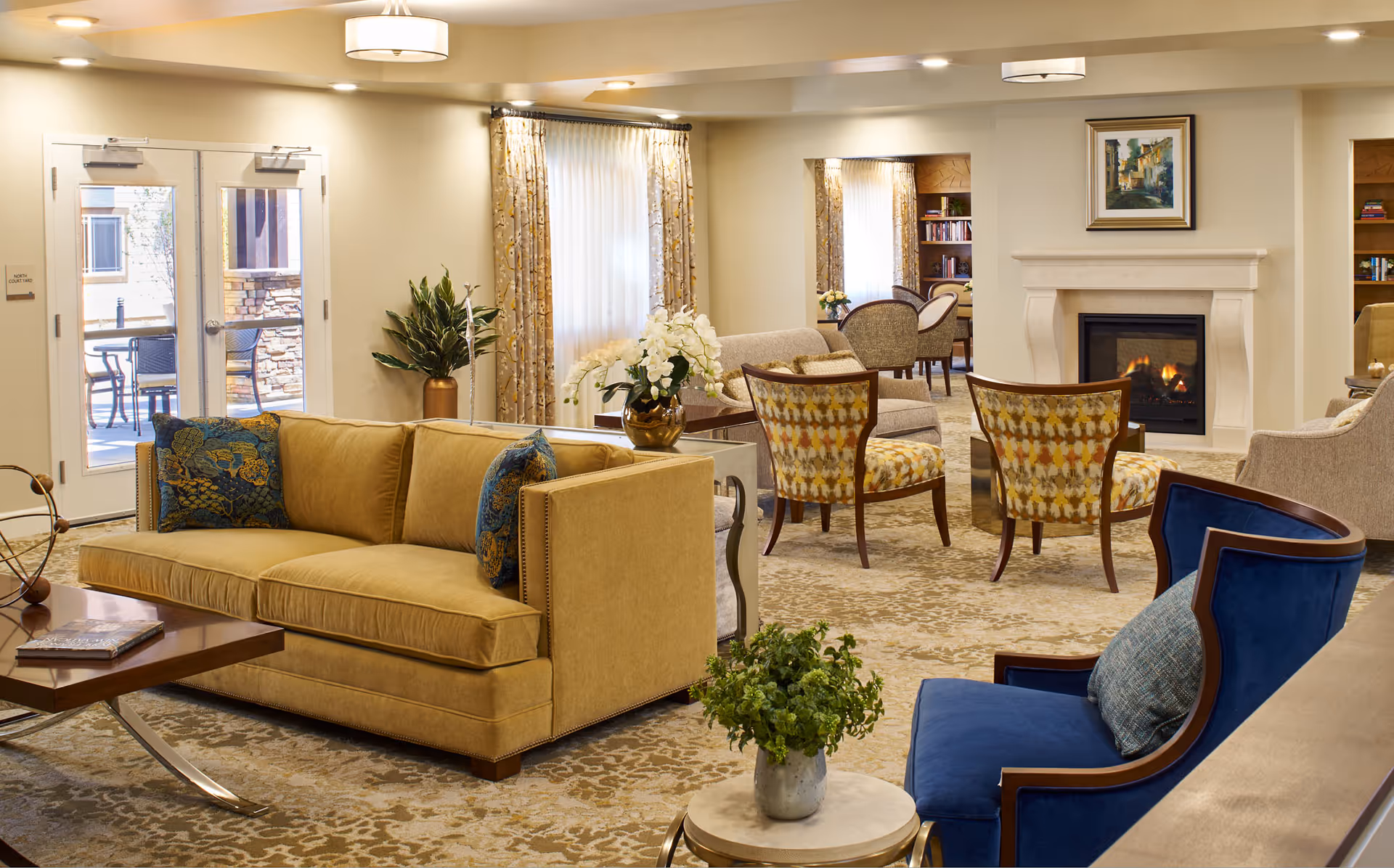 A cozy and well-lit living room area in a senior living facility with a beige sofa adorned with blue patterned pillows, a blue armchair with a cushion, and several patterned chairs arranged around a fireplace. The room features large windows with curtains, a coffee table with a book, decorative plants, and a warm, inviting atmosphere.