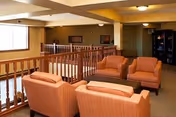 A cozy seating area in a senior living facility with four orange upholstered armchairs arranged around a small wooden coffee table. The space features a wooden railing overlooking a lower level, warm beige walls, and soft lighting from ceiling fixtures. There are framed pictures on the walls and a bookshelf in the background.