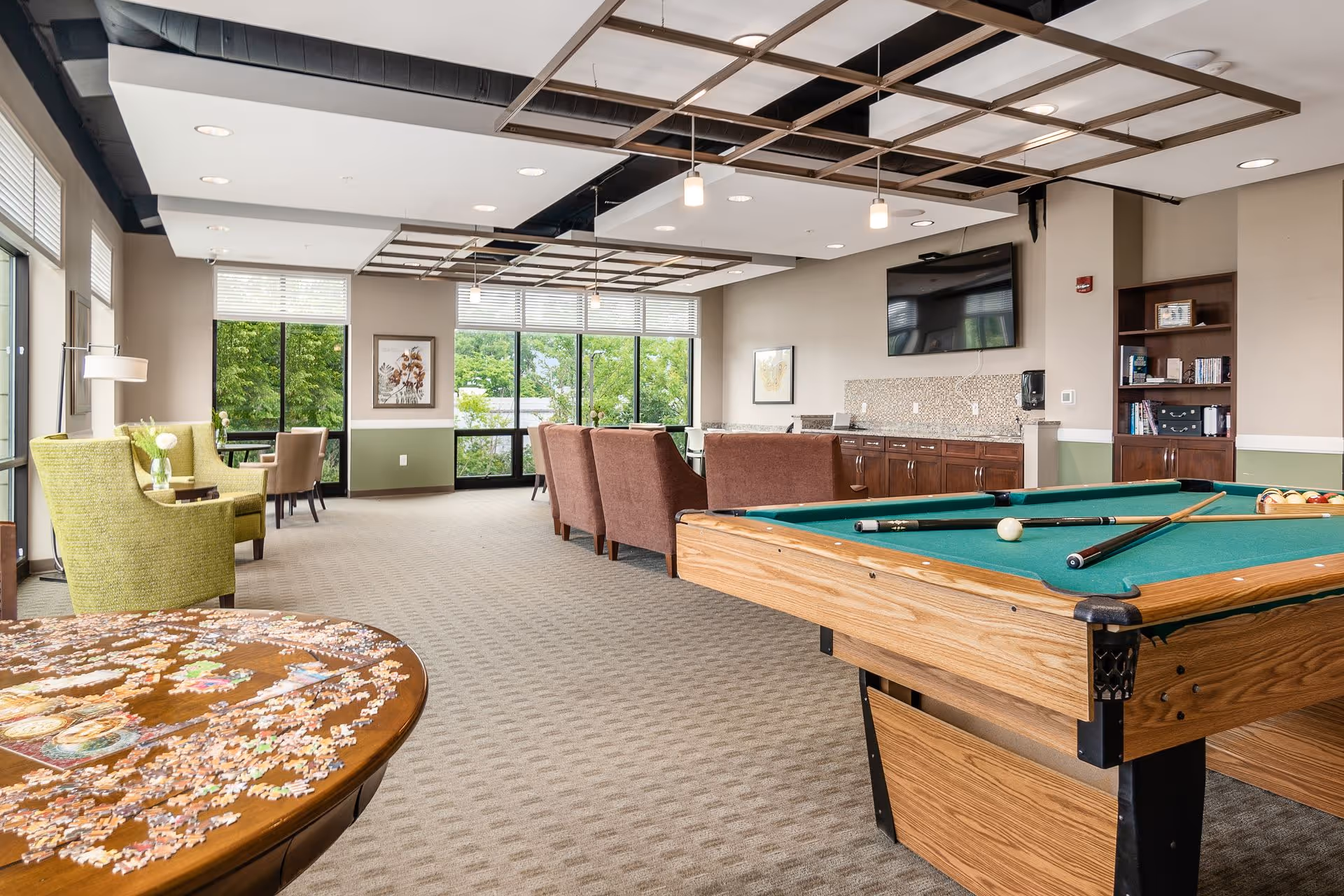 Bright communal activity room with a pool table, seating areas, a TV, and a puzzle table by large windows.